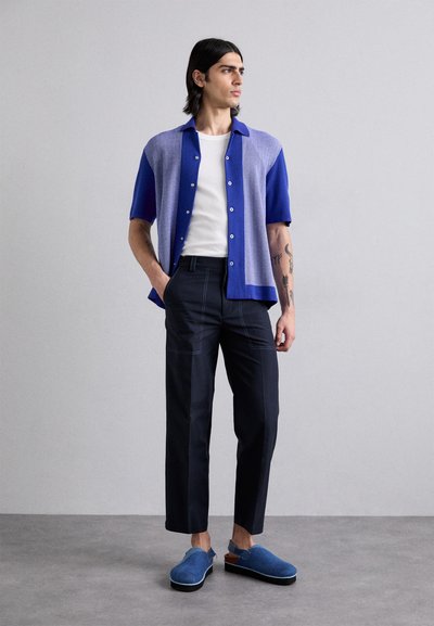 Short-sleeve blue and purple knit shirt with buttons, white tank top, navy trousers, and blue slip-on shoes. Clean lines and soft fabric.