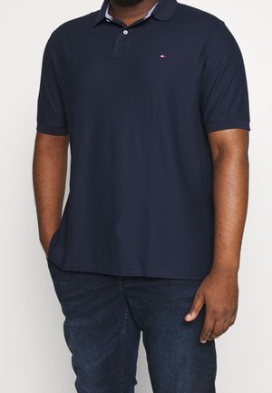 Mann trägt ein marineblaues Polohemd mit kurzen Ärmeln und einem kleinen Logo auf der Brust sowie dunkelblaue Jeans, steht mit einer Hand in der Hosentasche.