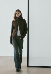 Młoda kobieta spacerująca w pomieszczeniu, ubrana w brązowy sweter rozpinany na zamek, biały top, ciemne szerokie dżinsy i czarne buty.