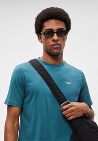 Tricou de culoarea albastru-verzui, cu gât rotund, din material moale, decorat cu un logo alb mic. Modelul poartă ochelari de soare negri și o geantă de umăr neagră.