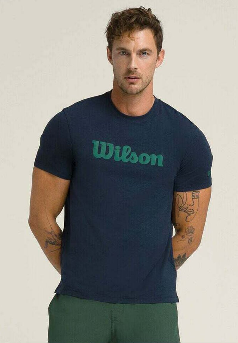 Camiseta de algodón azul marino de manga corta, con un logotipo verde de "Wilson" en una fuente curva en el pecho. Usuario lleva pantalones verdes.