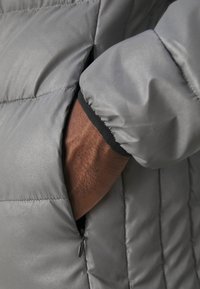 Graue Steppjacke mit einem Reißverschlussfach. Das Material ist glatt mit einer glänzenden Oberfläche. Bündchenärmel mit einem kontrastierenden schwarzen Saum.