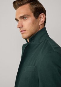 Veste vert foncé avec col montant, dotée d'une poche poitrine unique et d'un tissu texturé. Lignes épurées et design minimaliste.