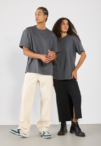 To personer står side om side. Den venstre bærer en løs grå t-shirt og cremefarvede bukser; den højre har en grå t-shirt og en sort nederdel med slids.
