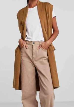Personne portant un t-shirt blanc, un pantalon beige taille haute et un gilet marron sans manches, debout les mains dans les poches.
