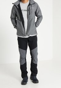 Veste zippée grise, t-shirt gris avec logo, pantalon noir et gris avec accents en panneaux, porté avec des chaussures de randonnée bleues sur fond uni.