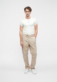 Balta piegāzta T-kreklu ar mazu melnu logotipu, kombinēta ar bežām brīva piegriezuma biksēm ar sānu kabatām un pogu aizdari. Baltas kedas.