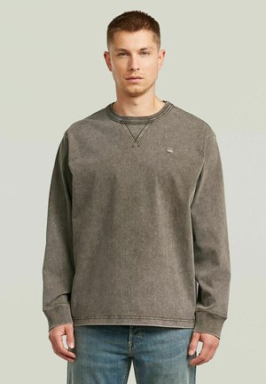 Giovane uomo in piedi, rivolto verso l'obiettivo, con una felpa grigia a maniche lunghe e jeans blu, su uno sfondo semplice.