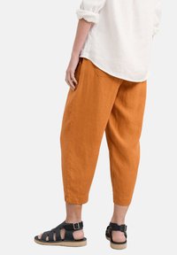 Rust-colored linen pants with a relaxed fit, cuffed hems, and back pockets, paired with black strappy sandals and a white shirt.