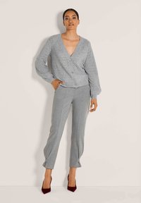 Vrouw in een lichtgrijze textuur cardigan met knopen, bijpassende grijze op maat gemaakte broek met enkelbandjes, en donkerrode hoge hakken.