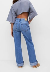 Jean bleu en denim taille haute avec une coupe décontractée, des coutures visibles et des revers retroussés. Le dos comporte deux poches et une étiquette en cuir marron clair.