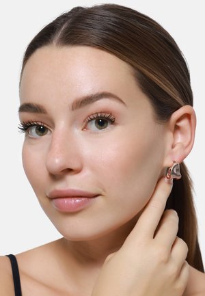 Roségoldene geometrische Ohrringe mit glänzender Oberfläche und geschwungenem Design, getragen von einem Model, das mit dem Finger einen Ohrring berührt.
