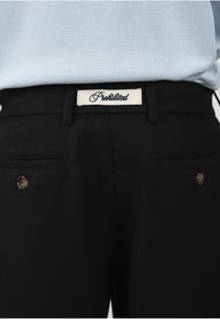 Gros plan d'un pantalon noir avec des poches latérales boutonnées et une étiquette blanche avec le texte en cursive « Privileged » à la taille, porté avec une chemise bleu clair texturée.