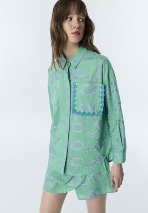 Femme portant une chemise ample verte à motif cachemire avec une poche bordée de bleu et un short assorti sur un fond clair uni.