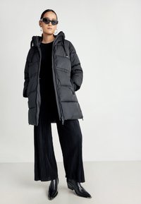 Svart puffer-jakke med hette, glidelås og quiltet design. Kombinert med svarte plisserte bukser og spisse ankelstøvler.