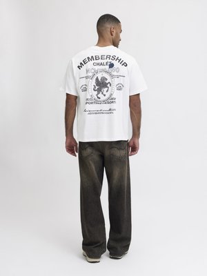 Homme de dos portant un T-shirt blanc imprimé avec des textes noirs et un blason de lion, assorti à un jean sombre ample et des baskets blanches.