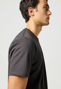 Camiseta de algodón gris oscuro con cuello redondo y mangas cortas. Costuras visibles a lo largo de los hombros y las costuras laterales. Textura suave.