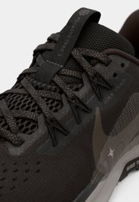 Baskets noires et brunes avec un dessus en maille et à motifs, bout renforcé, semelle texturée et accents réfléchissants. Les lacets s'entrelacent de manière sécurisée.