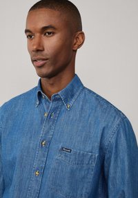 Denim overhemden in medium blauw met een borstzak, puntige kraag en gouden hardware details op de knopen. Zachte textuur.