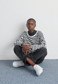 Svart-hvitt stripete langermet topp, svarte løse bukser og hvite joggesko. Modell sitter med hendene på knærne. Enfarget bakgrunn.