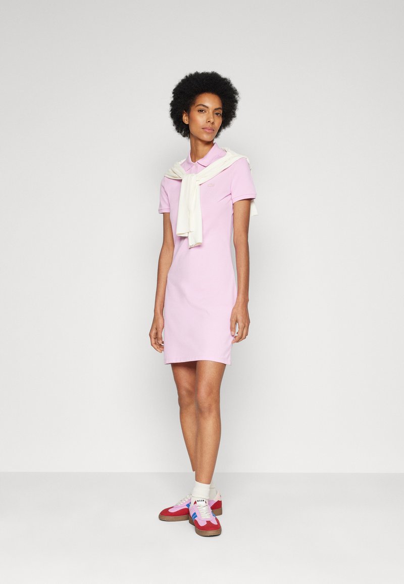 Vestido estilo polo en tela de color rosa claro con cuello, combinado con un suéter crema colocado sobre los hombros y zapatillas deportivas coloridas.