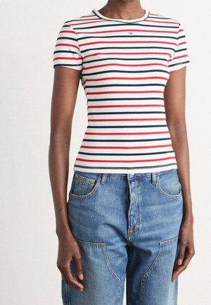 Osoba ubrana w białą koszulkę z krótkim rękawem z czerwonymi i granatowymi poziomymi paskami oraz niebieskie, wysokiej talii dżinsy.