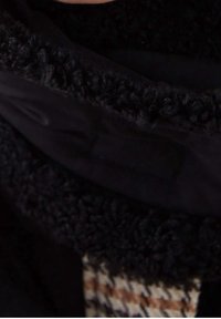 Primer plano del cuello de una chaqueta de forro polar negra con una tela texturizada y un destello de ropa con un patrón de ajedrez marrón y crema por debajo.