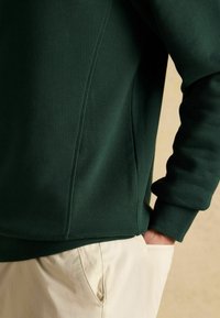 Osoba w ciemnozielonym swetrze z żebrowanymi detalami i jasnobeżowych spodniach, z ręką swobodnie w kieszeni na neutralnym tle.