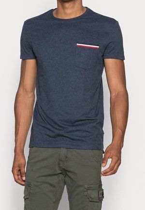 Hombre con una camiseta de manga corta azul marino con un pequeño bolsillo en el pecho que tiene ribetes rojos, blancos y azul marino, combinada con pantalones cargo verde oliva.