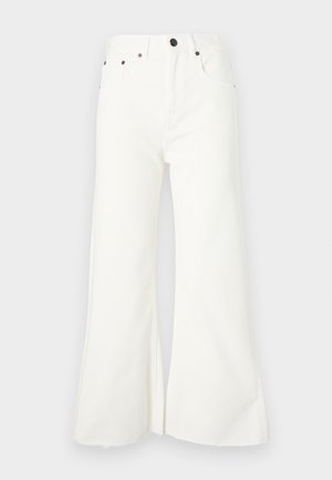 Suéter beige de media manga con cuello de cremallera, combinado con jeans anchos blancos. La modelo lleva un clutch negro y calza zapatos de punta negros.