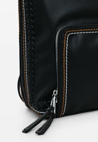 Detailní záběr rohu černé kožené tašky s pleteným lemováním, stříbrným zipem a oranžovým a bílým prošíváním na okraji kapsy.