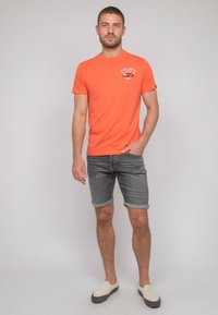 T-shirt orange à manches courtes avec un graphique sur le devant, associé à un short en denim gris et des chaussures légères à enfiler. Fond uni.