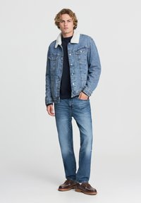 Uomo in piedi indossa una giacca di jeans foderata in sherpa blu, un maglione scuro, jeans blu e scarpe stringate in pelle marrone su uno sfondo bianco semplice.