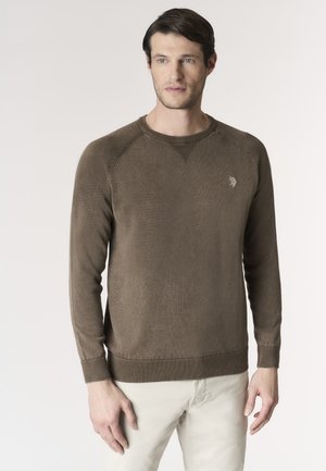 Uomo con capelli corti e scuri che indossa un maglione marrone a maniche lunghe e pantaloni beige chiaro, in piedi davanti a uno sfondo chiaro e uniforme.