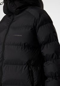 Svart pufferjacka med hög krage, dragkedjestängning och synlig kviltad sömnad. Har en logotyp på bröstet och sidofickor.