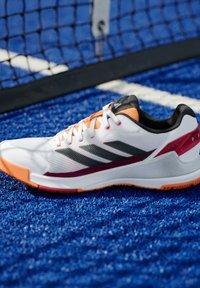 Witte sportschoen met zwarte en grijze strepen, oranje accenten en een gestructureerde mesh-bovenkant. Voorzien van een rubberen zool voor grip op blauw kunstgras.