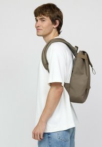 Bruine stoffen rugzak met een rolsluiting, gevoerde banden en een gestructureerd oppervlak, gedragen door een persoon in een wit shirt en blauwe jeans.