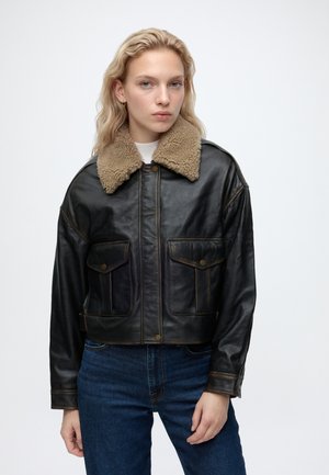 Jonge vrouw met golvend blond haar die een zwarte leren jas met een beige shearlingkraag en blauwe spijkerbroek draagt, staand tegen een effen achtergrond.