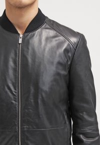 Zwarte leren bomberjack met geribbelde kraag, met gladde en geperforeerde texturen op de mouwen en voorkant. Ritssluiting.