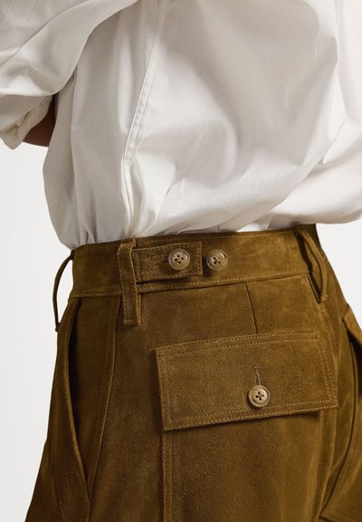Smeđe antilop hlače s dvostrukim kopčama na gumbu, bočnim džepovima i zakrpnim džepom straga, uparene s bijelom košuljom sa šlicem na gumbe.