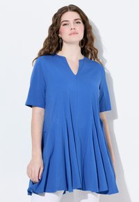 Túnica corta de manga corta azul con escote en V, diseño plisado y tela suave y lisa, combinada con pantalones blancos.