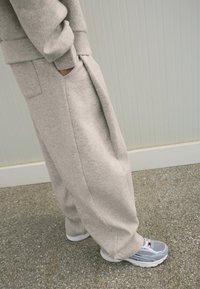 Osoba stojící s rukama v kapsách, oblečená v světle šedých oversized teplácích a odpovídajícím svetru, s šedobílými běžeckými botami.
