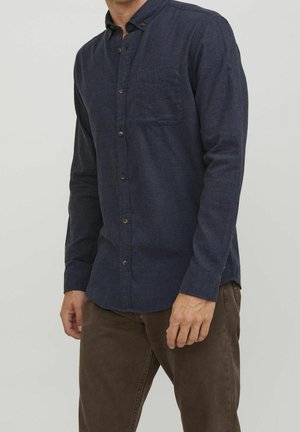 Homme portant une chemise bleu foncé boutonnée avec une poche poitrine et un pantalon marron, debout devant un fond clair uni.