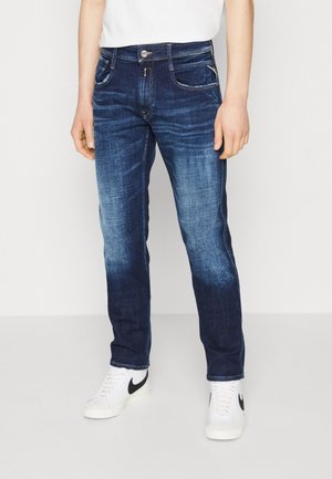 Vaqueros de mezclilla azul oscuro con un diseño desgastado, cinco bolsillos, costuras en contraste y cierre de botón. Combinados con zapatillas blancas.
