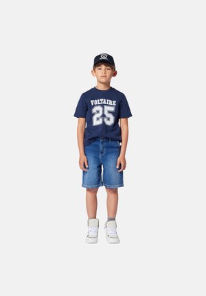 T-shirt in cotone navy con la scritta "VOLTAIRE 25" in bianco, abbinata a pantaloni corti in denim e sneaker alte bianche.