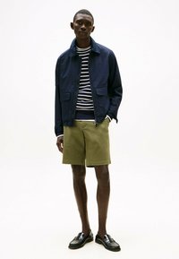 Giacca leggera blu navy, camicia a maniche lunghe a righe, pantaloni corti verde oliva e mocassini neri. Texture morbide, design semplice e colori a contrasto.