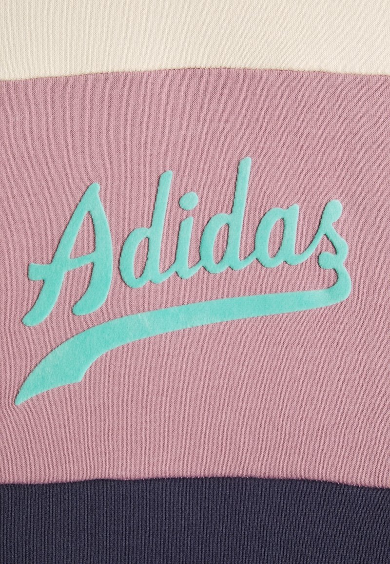 Felpa Adidas con un corpo rosa chiaro e il logo turchese ricamato su un tessuto morbido, bordato di un blu navy scuro nella parte inferiore.