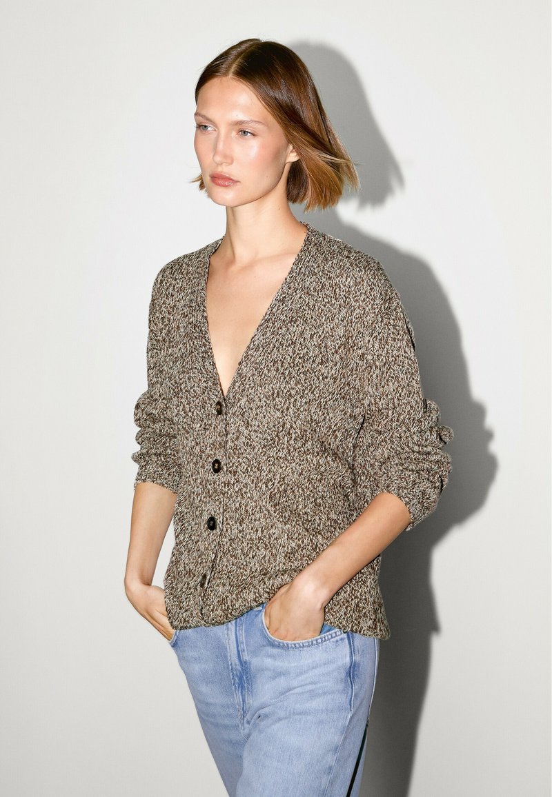 Cardigan lavorato a maglia in tonalità marroni terrose con scollo a V profondo, cinque bottoni e maniche arrotolate, abbinato a jeans in denim azzurro chiaro.