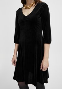 Schwarzes Samtkleid mit V-Ausschnitt, dreiviertel langen Ärmeln und einem leicht gerafften Detail an der Brust. Ausgestellter Rock und glatte Textur.