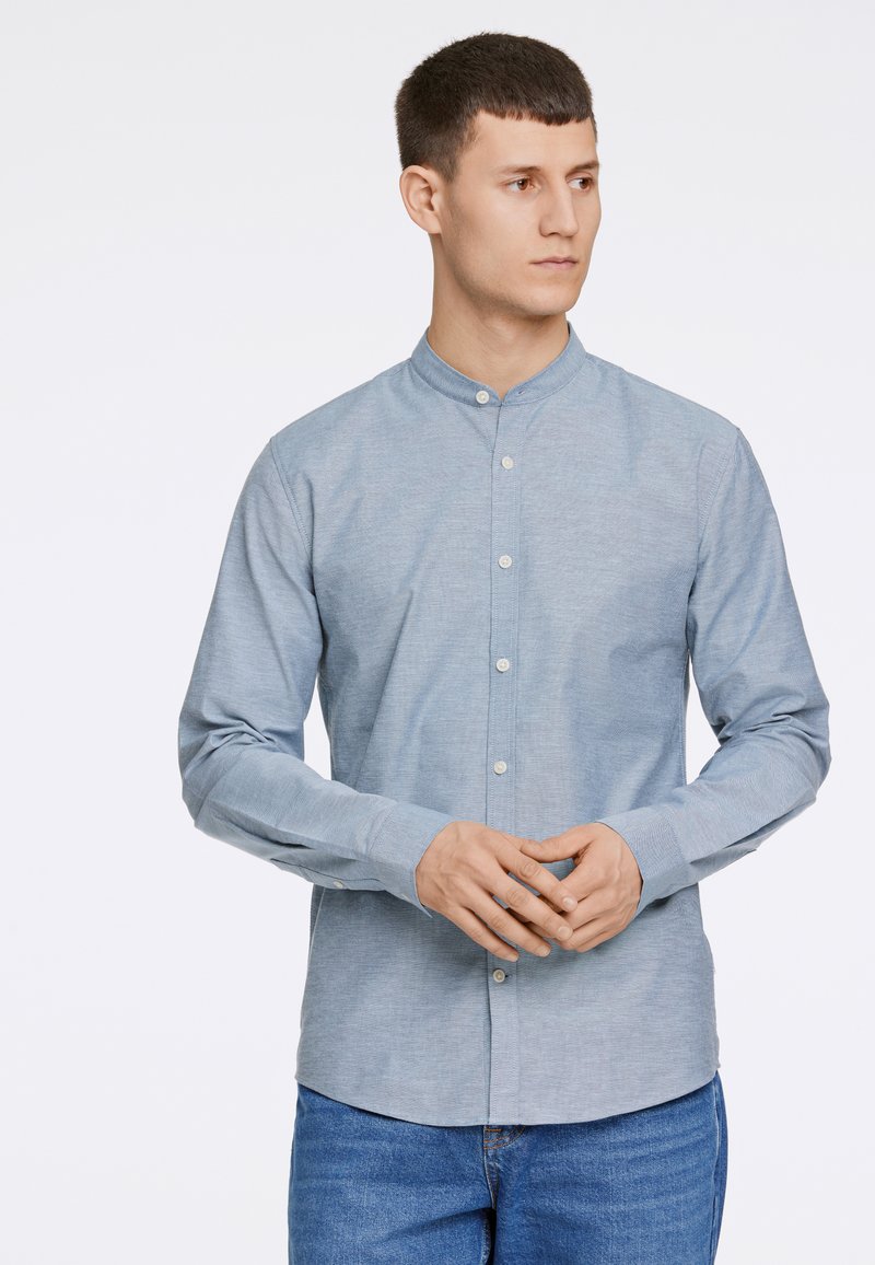 Camisa de botón celeste con cuello mandarina, mangas largas, un bolsillo en el pecho y una textura sutil. Llevada por una persona.